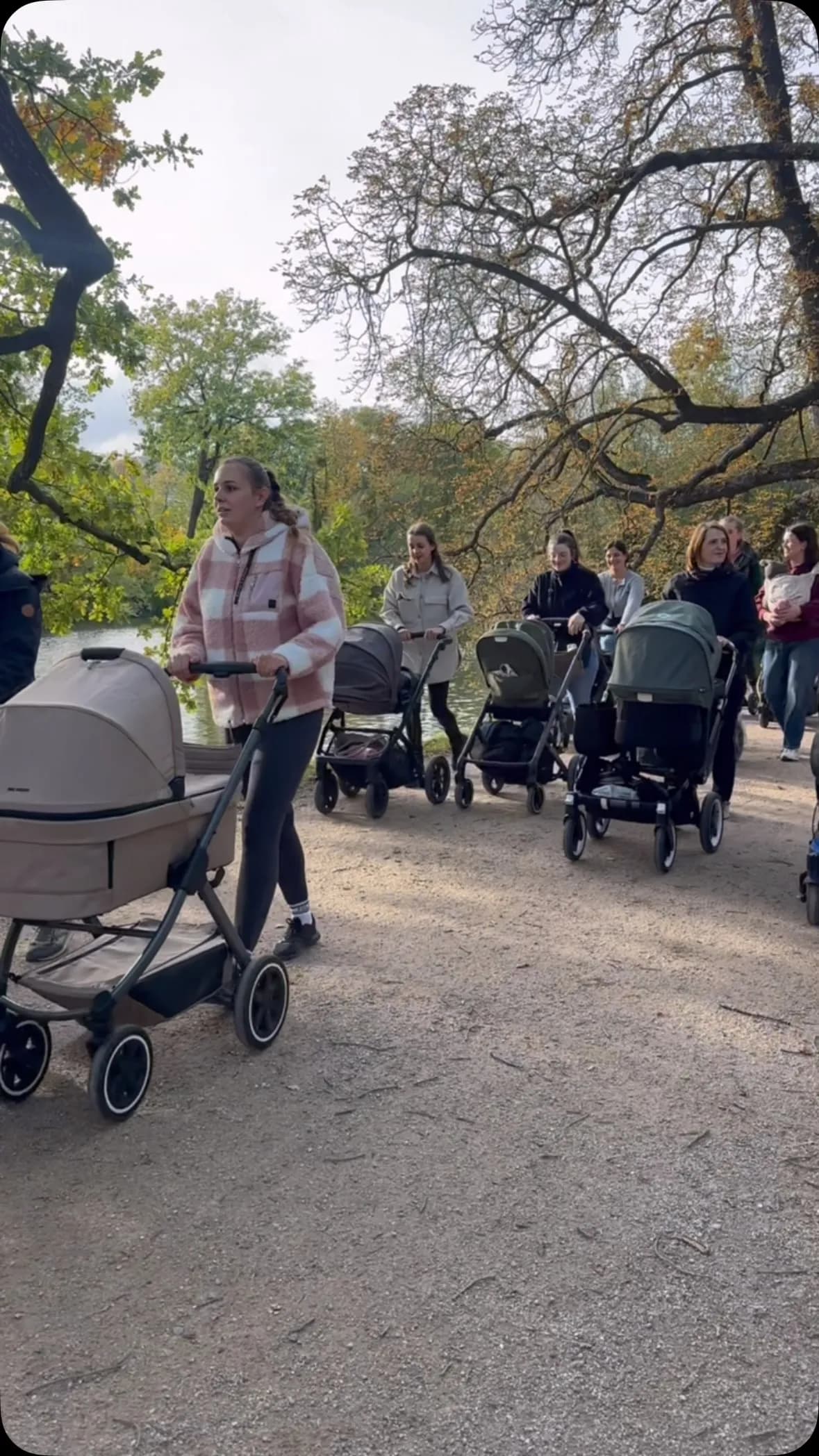Mamas schieben Kinderwagen auf einem Waldweg im Herbst, entlang eines Sees mit bunten Bäumen.