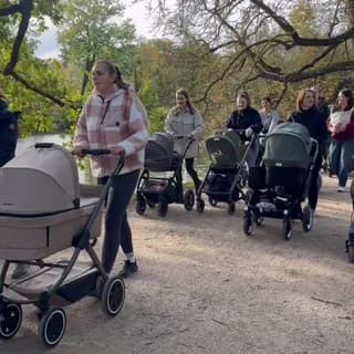 Mamas schieben Kinderwagen auf einem Waldweg im Herbst, entlang eines Sees mit bunten Bäumen.