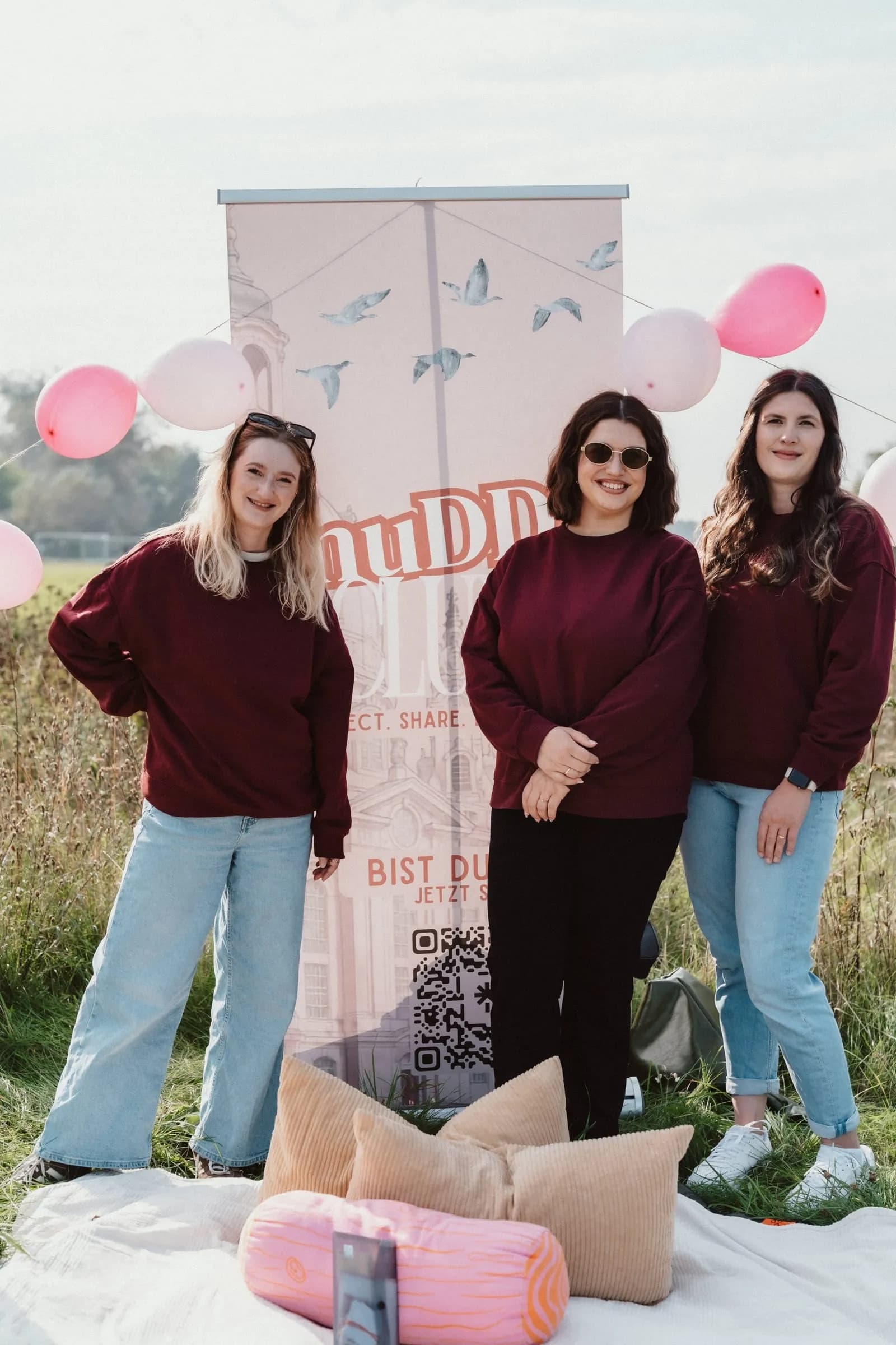 Das muDDi Club Team lacht beim Picknick vor dem Banner des Clubs, daneben rosa Luftballons